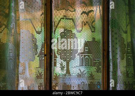 Vintage white openwork transparent curtains with houses, trees, plants, flowers, birds, clouds pattern in front of the light on the old wooden window Stock Photo