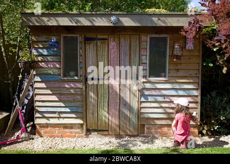 Young girl sat on floor colouring in shed wall in garden, Uk Stock Photo