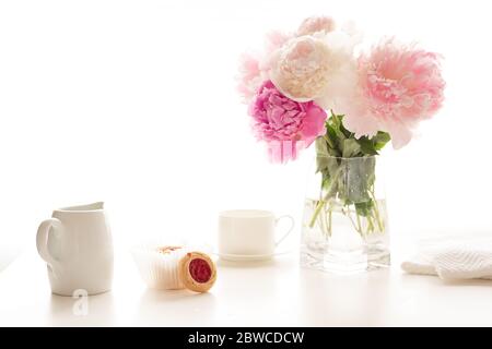 Pink peonies in glass vase, cup of tea, and cookies close up on white kitchen table in morning sunshine. Romantic breakfast, love, wedding, celebratio Stock Photo