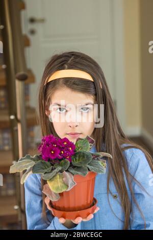 The girl is sitting in the kitchen and shows a beautiful flower in a pot Stock Photo