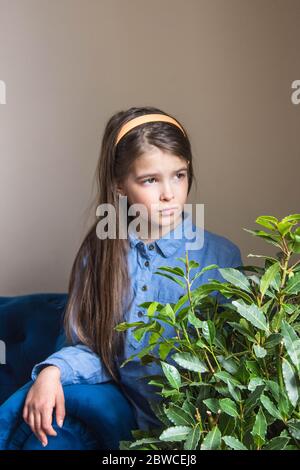 A girl in a denim dress sits and looks out the window in a room with a blue sofa and a flower Stock Photo