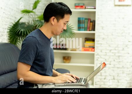 Young smiling Asian man in casual clothing on sofa working on laptop computer. Male freelancer working from home on notebook. Work from home concept Stock Photo