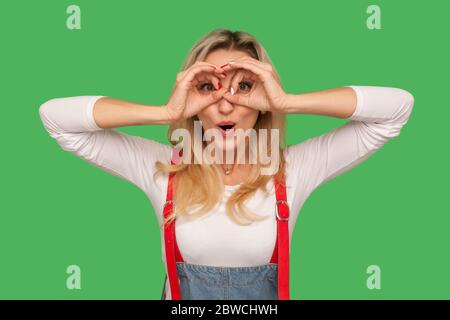 Portrait of curious amazed adult woman in denim overalls standing with binoculars gesture, looking surprised to discover and observe shocking content. Stock Photo