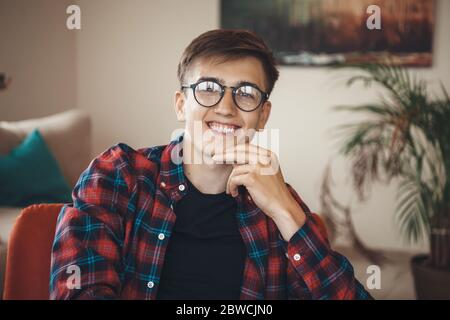 Young caucasian man with eyeglasses wearing a shirt is posing on armchair touching his chin and smile at camera Stock Photo