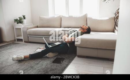 Tired caucasian man falling asleep on the floor with a computer and tablet with crossed hands Stock Photo