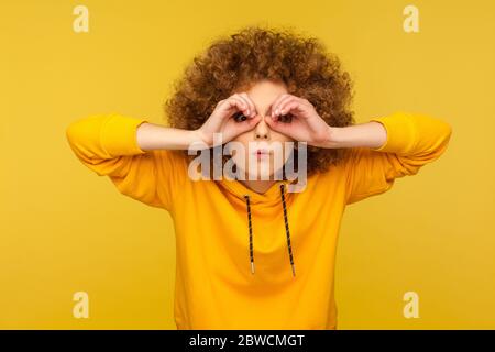 What is there? Portrait of surprised curly-haired woman in urban style hoodie making glasses shape, looking through binoculars gesture with shocked ex Stock Photo