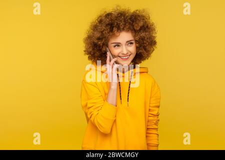 Portrait of cheerful curly-haired woman in urban style hoodie calling friend, talking carefree on smartphone and smiling joyfully, discussing great ne Stock Photo