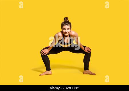 Motivated assertive gymnast girl with hair bun in tight sportswear squatting and spreading legs to stretch muscles, warming up before twine exercise, Stock Photo