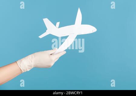 Profile side view closeup of human hand in white surgical gloves holding airplane paper. indoor, studio shot, isolated on blue background. Stock Photo