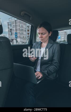 Businesswoman using mobile phone and laptop computer in car whole sitting at the back seat, business communication on the move Stock Photo