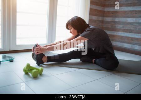 Adult fit slim woman has workout at home. Senior model sit with crossed legs and meditating or praying. Stretching hands holding together. Take care Stock Photo