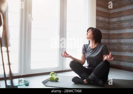 Adult fit slim woman has workout at home. Process of senior model meditating. Hold hands on knees and look at window. Stock Photo
