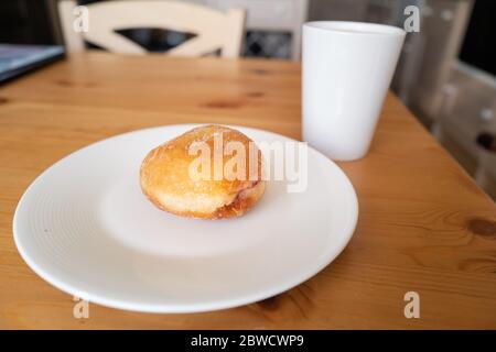 A jam doughnut on a white plate next to a white mug cup on a wood kitchen table with a wooden chair. Soft focus Stock Photo