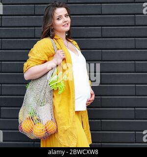 Pregnant woman in yellow clothes with an eco bag with fruits and vegetables on a black wall background. Plastic free concept Stock Photo