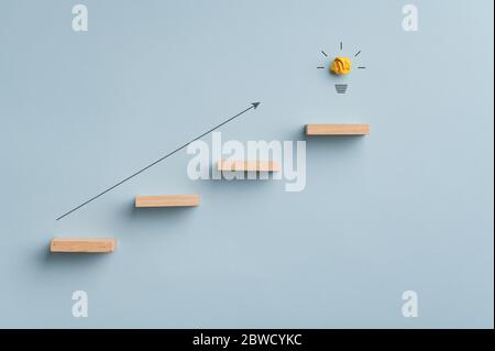 Arrow pointing up the staircase made of wooden pegs with glowing light bulb on the top one. Conceptual image of idea, innovation and ambition. Stock Photo