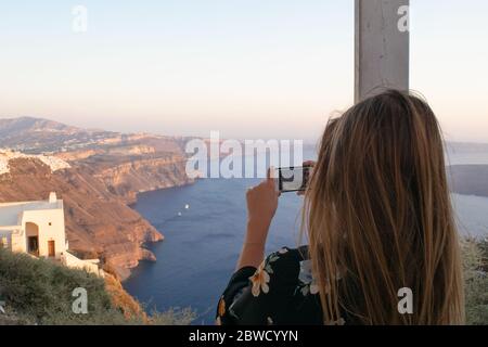 Blonde girl in flower summer dress takes photo of the stunning view of the cliffs of Santorini Stock Photo