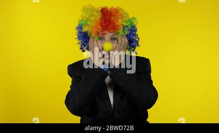 Senior caucasian female clown in colorful wig and blue bow tie looks surprised shocked. Amazement. Funny elderly woman in Halloween costume. People emotions. Copy space. Yellow background. Chroma key Stock Photo