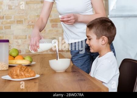 Selective focus of mother pouring milk in bowl with cereals near smiling son in kitchen Stock Photo