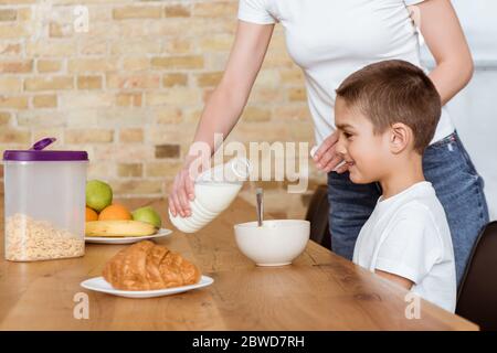 Mother pouring milk in bowl with cereals near smiling son at kitchen table Stock Photo