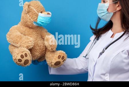 woman doctor pediatrician holds brown teddy bear in hand in white medical disposable mask, concept of preventing epidemics and pandemics against flu Stock Photo
