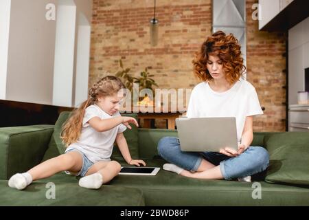 redhead freelancer using laptop near upset daughter with digital tablet Stock Photo