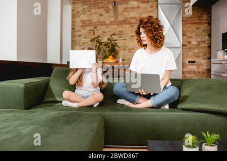 redhead freelancer using laptop near daughter covering face with digital tablet Stock Photo