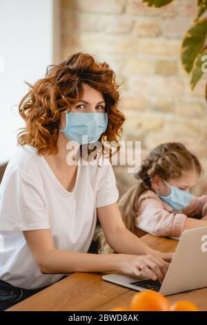 selective focus of redhead freelancer mother in medical mask using laptop near daughter Stock Photo