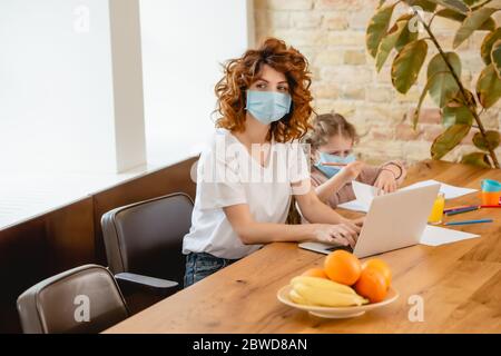 selective focus of curly freelancer mother in medical mask using laptop near daughter Stock Photo