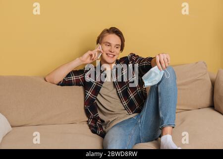 handsome holding medical mask and smiling while talking on smartphone in living room Stock Photo
