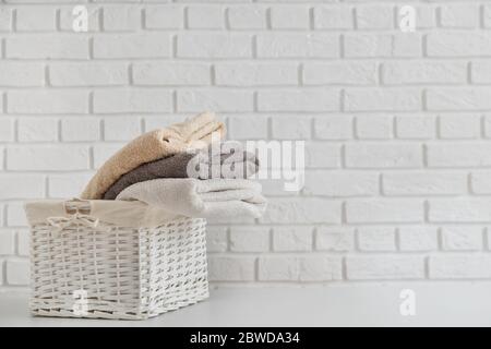 Multicolor fresh towels in wicker straw basket on white table with white brick wall on backdrop. Stock Photo