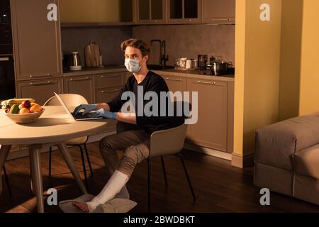 Side view of man in medical mask and plaster bandage on led using laptop in kitchen Stock Photo