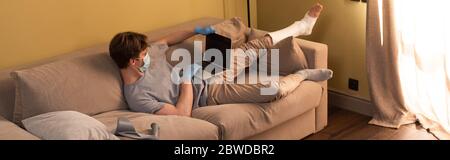 Panoramic concept of freelancer in medical mask with broken leg working on laptop at home Stock Photo