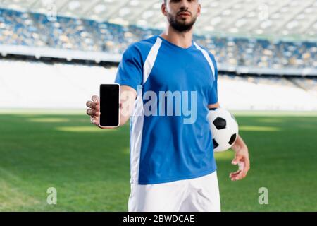 cropped view of professional soccer player in blue and white uniform with ball showing smartphone with blank screen at stadium Stock Photo