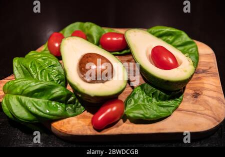 A wooden cutting board on a black background with an avocado in the center. All around, intense green basil leaves and bright red cherry tomatoes. Stock Photo