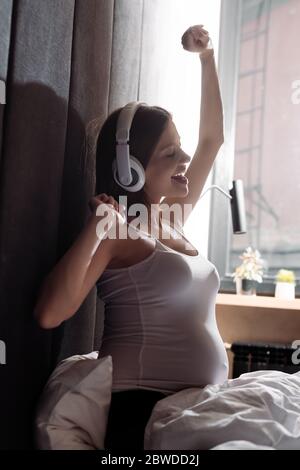 happy and pregnant woman with closed eyes and wireless headphones  listening music in bedroom Stock Photo