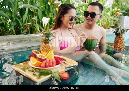 Happy young couple drinking cocktails from small watermelon and pineapple and eating fresh fruits when resting in small swimming pool of spa hotel Stock Photo
