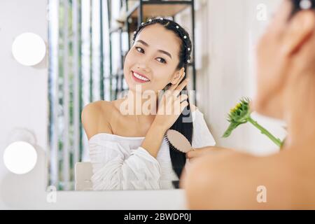 Smiling charming young Vietnamese woman sitting at her vanity and brushing hair Stock Photo