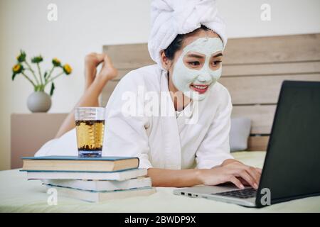 Cheerful female student lying in bed with glass of green tea on stack of books and watching webinar Stock Photo