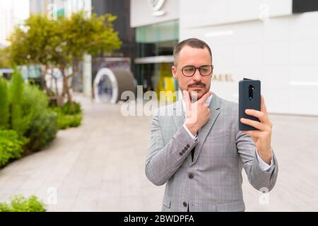 Handsome Hispanic bald bearded businessman taking selfie in the city outdoors Stock Photo