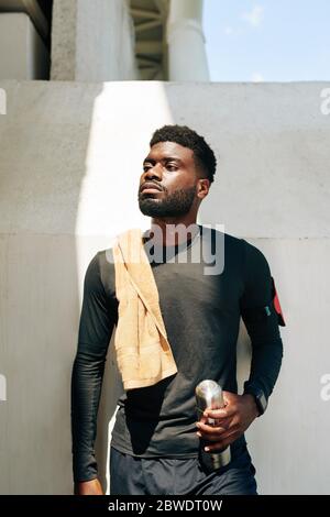 Portrait of serious confident young sportsman with towel on his shoulder standing with thermos in hand and looking away Stock Photo