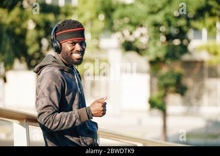 Portrait of positive young Black man listening to music in his headphones and showing thumbs-up Stock Photo
