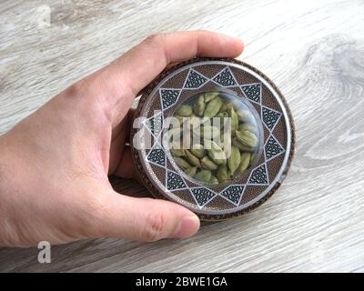 mashhad, IRAN, 01 03 2020: Green cardamom pods into handcraft box on wooden background. Hand on box. Close up. Stock Photo