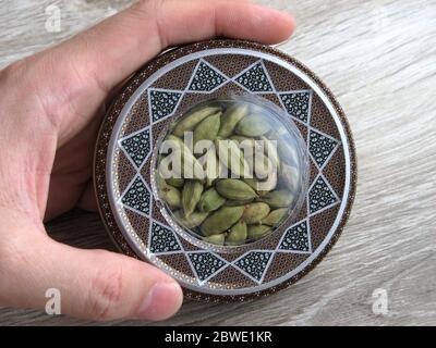 Mashhad, IRAN, 01 03 2020: Close up of green cardamom pods into handcraft box on wooden background. Hand on box. Stock Photo