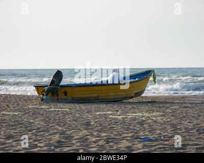 Old yellow  wooden fishing boat with fishing man aground on the beach, near sea caspian, Iran. A man sitting on a motor boat on the seashore. Stock Photo