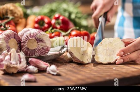 Detail of garlic head being cut to half with more fresh unpeeled garlic on the side and various veg in the background Stock Photo