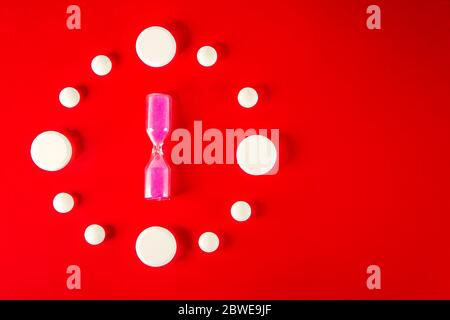 Clock made of white tablets with hourglass in the middle on red background, top view Stock Photo