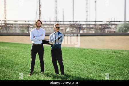 Two young engineers standing outdoors by oil refinery, discussing issues. Stock Photo