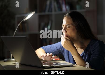 Tired woman with laptop yawning sitting late with a desk lamp on the table at night at home Stock Photo