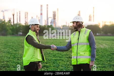 Two young engineers standing outdoors by oil refinery, shaking hands. Stock Photo