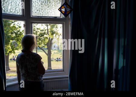 With the UK death toll reaching 38,161, a further 324 victims in the last 24hrs, and the government's pandemic lockdown still in effect, a middle-aged woman looks out from a first floor window in deep thought, while viewing families and friends outside in a local park, on 29th May 2020, in London, England. Stock Photo